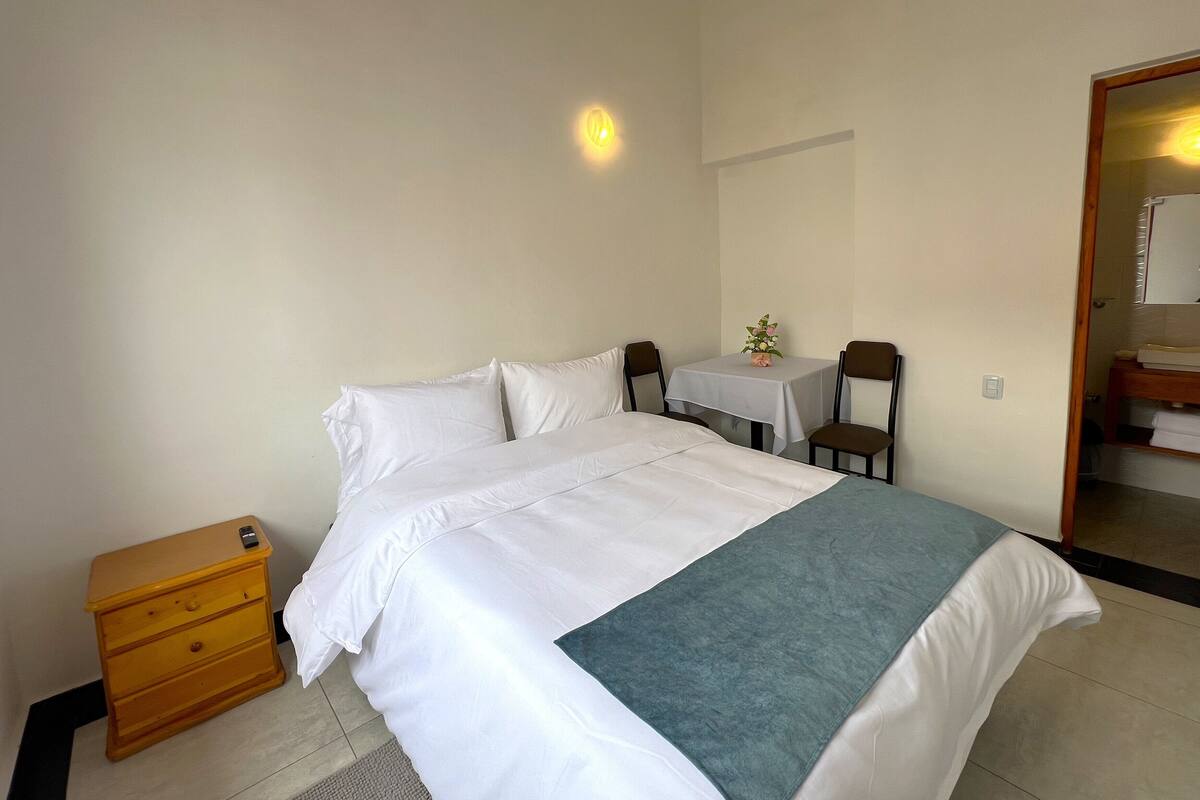 A neatly made bed with a white comforter and a teal blanket, a wooden nightstand with drawers, a small table with a vase of flowers, and a chair.