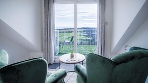 A room with a view of a green landscape through large windows, a small round table with a plant, and two green armchairs.