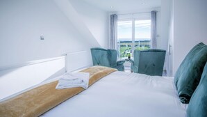 A hotel room with a bed, two chairs, a small table, and a view of a green landscape through large windows.