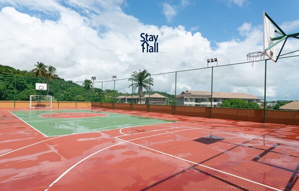 Una cancha de baloncesto al aire libre con superficie verde y roja, rodeada por una valla y un aro de baloncesto.