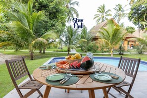 Mesa de comedor al aire libre con piscina al fondo, rodeada de palmeras.