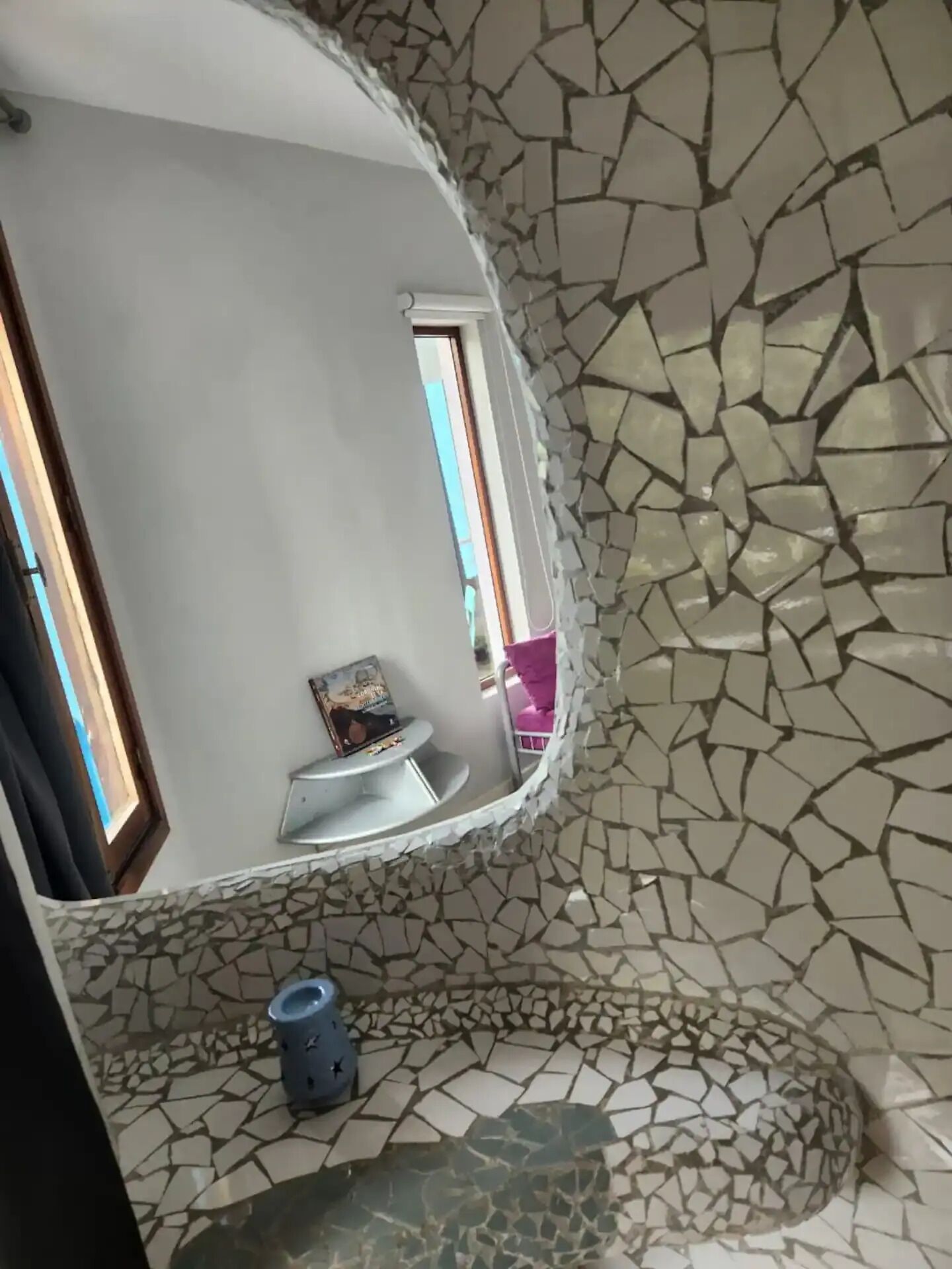 A curved room with a mosaic wall, a small seating area with a table and a chair, and a window.