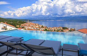 Une vue sur la piscine, avec des chaises longues, une vue dégagée sur une ville côtière et une étendue d’eau en arrière-plan.