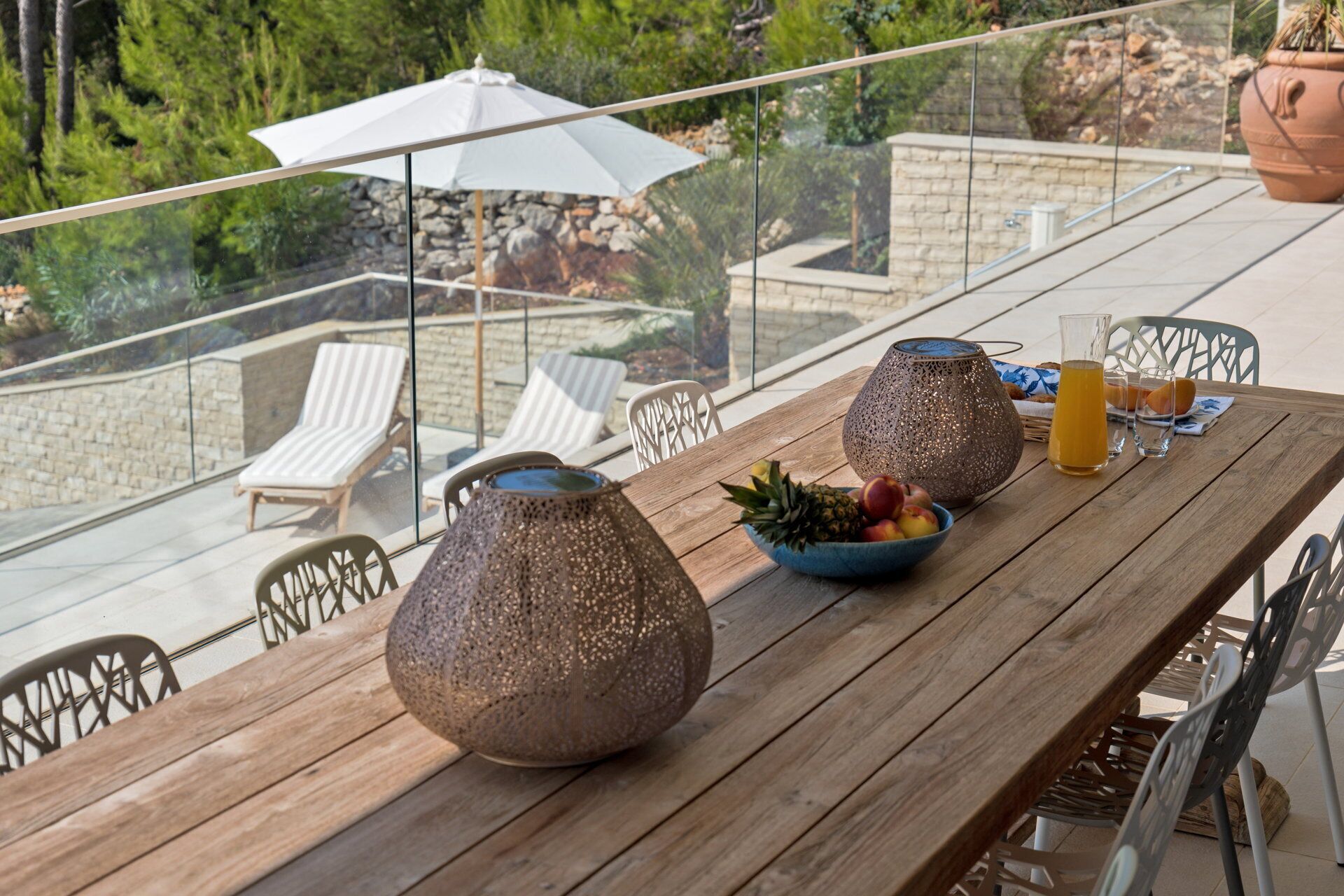A wooden table with two decorative vases, a bowl of fruit, and a glass of orange juice on a balcony with a glass railing, overlooking a garden and lounge chairs.