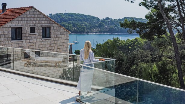 A woman standing on a balcony overlooking a body of water with boats, a stone building, and a forested hill in the background. - Villa Hestia with Heated Pool (Lumbarda)