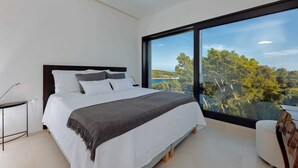 A modern bedroom with a large bed, a bedside table, and a view of a forest and sea through a large window. - Villa Hestia with Heated Pool (Lumbarda)