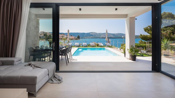 Un salon moderne doté d’une grande porte-fenêtre coulissante qui donne sur un espace piscine offrant une vue sur la mer et les montagnes.