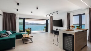 Un salon moderne avec un canapé vert, une table basse et un téléviseur. Une grande fenêtre offre une vue sur la mer.