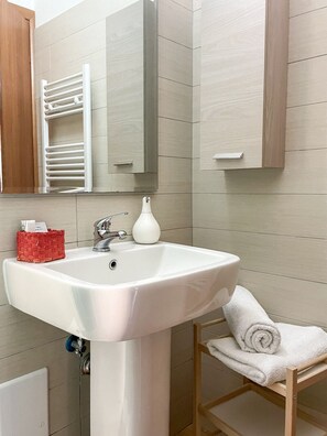 Un baño con un lavabo blanco, una cesta roja, un jarrón blanco y una silla de madera con una toalla.