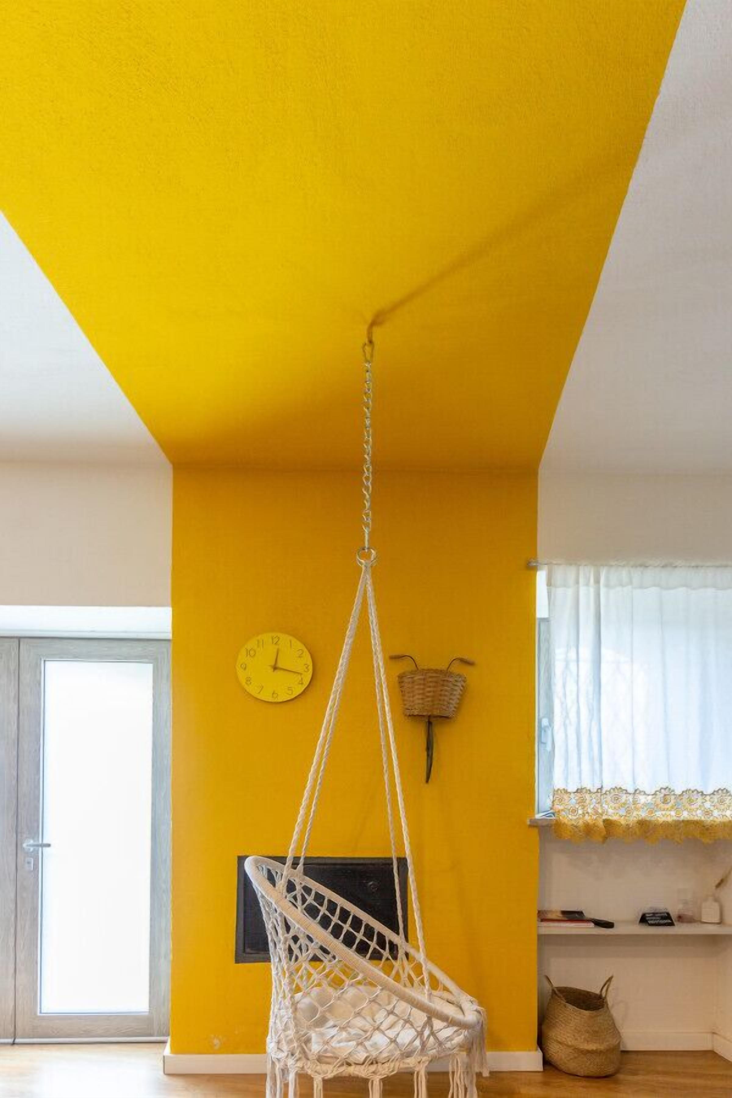A room with a yellow wall, a white hanging chair, a clock, and a window with curtains.