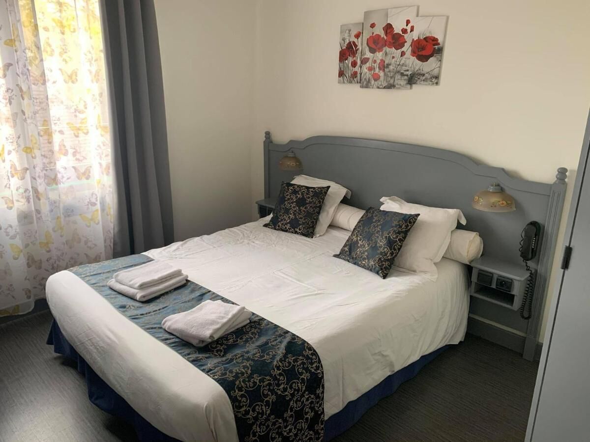 Een net opgemaakt bed met een wit-blauwe spreideken, twee kussens en opgevouwen handdoeken. Er staat een nachtkastje met een lamp en een telefoon.