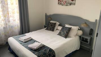 Een net opgemaakt bed met een wit-blauwe spreideken, twee kussens en opgevouwen handdoeken. Er staat een nachtkastje met een lamp en een telefoon.
