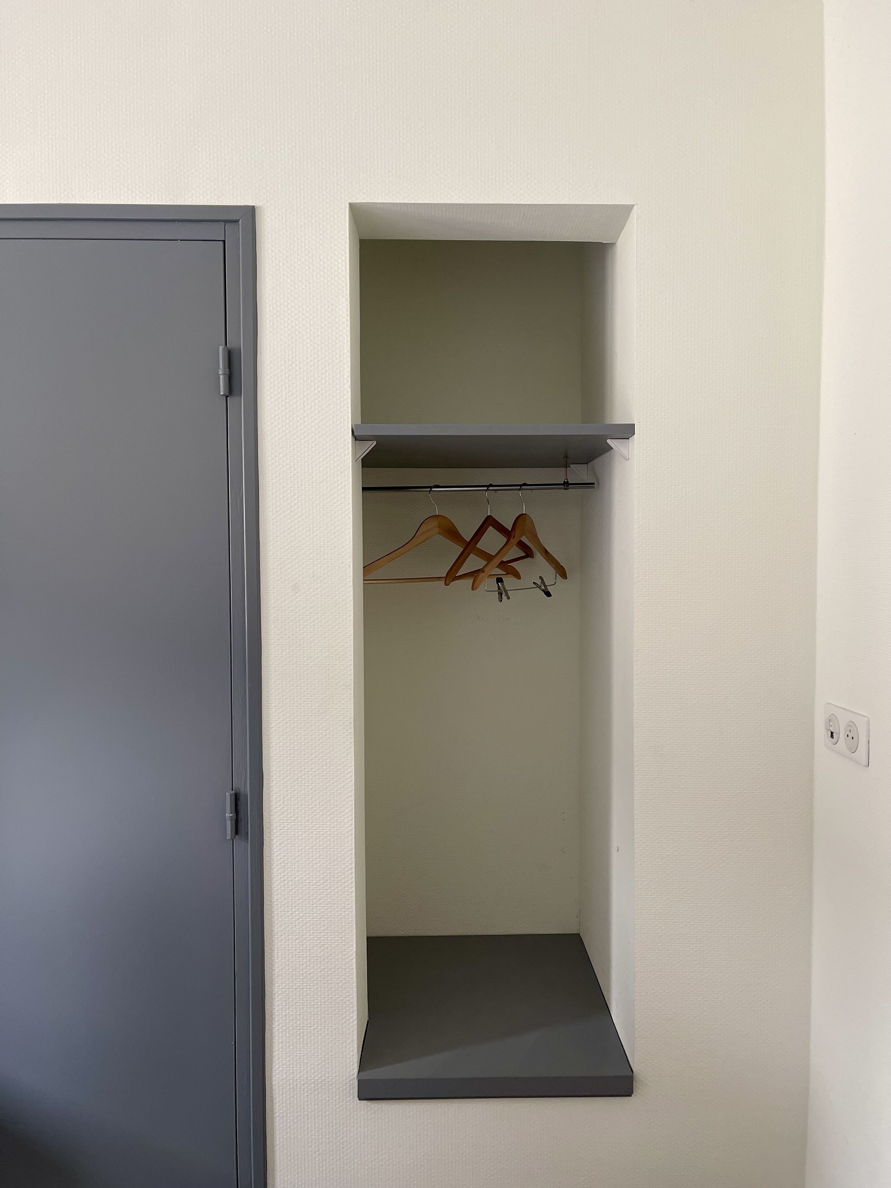 Een kamer met een grijze deur, een witte structuurwand, een zwarte plankkast met een hangstang, en een stopcontact.