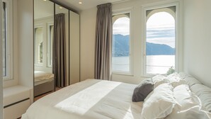 A bedroom with a large bed, a mirror, and two windows offering a view of a lake and mountains. - Villa Moltrasio - Four Bedroom Villa, Sleeps 8 (Moltrasio)