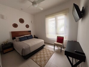 A bedroom with a bed, a desk, a chair, a TV, and a window with blinds.