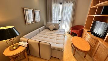 A hotel room with a large bed, a desk with a lamp, a TV on a wooden cabinet, and a window with curtains.