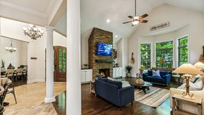 A spacious living room with a stone fireplace, a television mounted above it, and a large window with a view of trees. - Mountain Hideaway 8 acres - 5 Bedrooms + Hot Tub (Hendersonville)