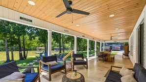 A covered outdoor seating area with wooden furniture, a ceiling fan, and large windows overlooking a grassy area. - Urban Rustic Lakeside Retreat with Dock, Sleeps 20 (Gallatin)