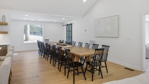 A modern dining room with a long wooden table and black chairs, white walls, and a large window. - Urban Rustic Lakeside Retreat with Dock, Sleeps 20 (Gallatin)