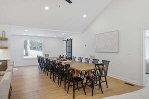 A spacious dining area with a long wooden table surrounded by chairs, a large window with a view of trees, and a ceiling fan.