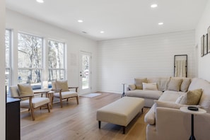 A spacious living room with a large sofa, two armchairs, and a coffee table. There are large windows providing natural light and a view of trees outside.