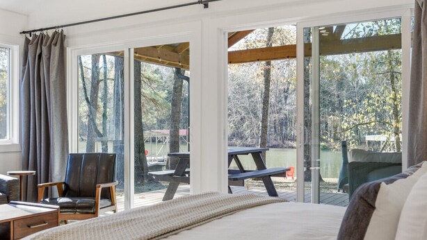 A bedroom with a large bed, a chair, a desk, and a view of a deck and outdoor area. - Urban Rustic Boathouse on Cove (Gallatin)