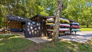 A wooden cabin with a covered porch, a small outdoor seating area, and a collection of kayaks and paddleboards stored on a rack. - Urban Rustic Boathouse on Cove (Gallatin)