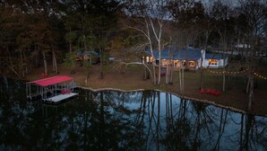 A lakeside property with a dock, a covered area with chairs, and a house with string lights. - Urban Rustic Boathouse on Cove (Gallatin)