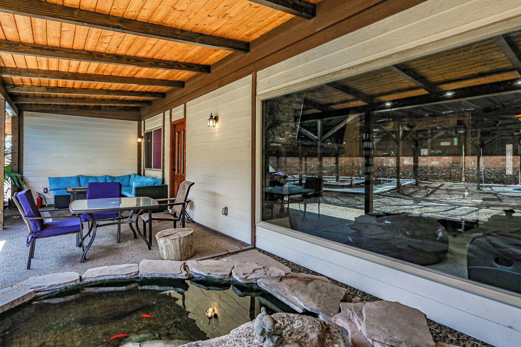 A covered outdoor seating area with a table and chairs, a small pond with fish, and a view of a parking lot through large windows.