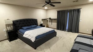 Un dormitorio con una cama grande, una mesita de noche, un ventilador de techo y una ventana con cortinas.