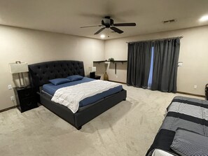 Un dormitorio con una cama grande, una mesita de noche, un ventilador de techo y una ventana con cortinas.