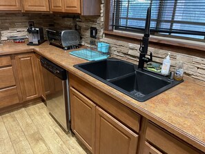 Una cocina con encimera de granito, pileta negra, armarios de madera, lavavajillas, tostadora y ventana con persianas.