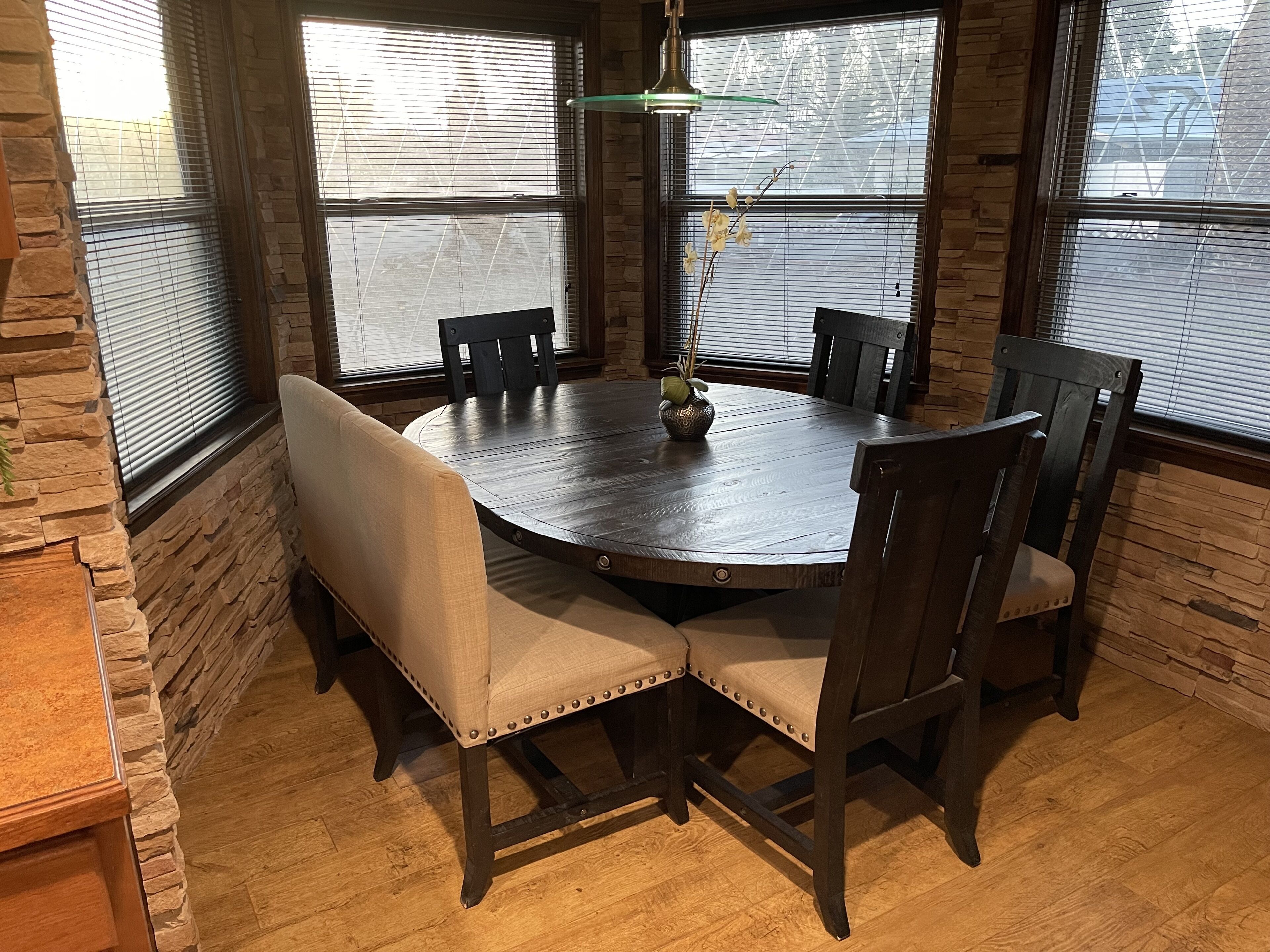 A round wooden dining table with six chairs, surrounded by stone walls and large windows with blinds.