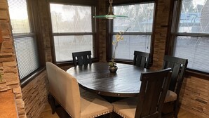 Una mesa de comedor redonda de madera con seis sillas, rodeada de paredes de piedra y grandes ventanales con persianas.