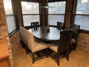 Una mesa de comedor redonda de madera con seis sillas, rodeada de paredes de piedra y grandes ventanales con persianas.