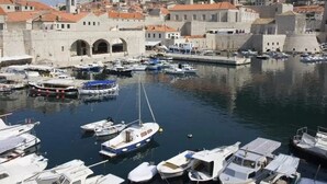 Un port de plaisance avec des bateaux amarrés, un bâtiment historique en pierre doté d’arcades, et une ville perchée sur une colline en arrière-plan.