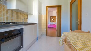 A compact kitchen with a stove, oven, and sink. A bed with a checkered bedspread is visible in the adjacent room. - Studio apartment with garden view balcony (Zadar)