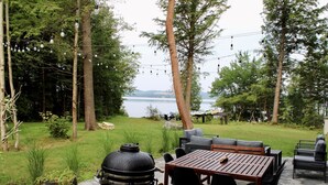 An outdoor patio with a wooden table and chairs, surrounded by greenery and a view of a lake. - Hôtel à la maison - Le refuge de Louise, avec spa (Weedon)