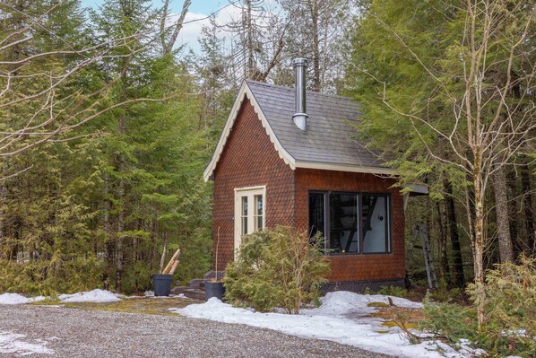 A small, rustic cabin with a sloped roof and a chimney, surrounded by dense forest.