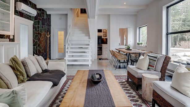 A modern living room with a sofa, coffee table, and a dining area with chairs and a table. A staircase leads to an upper level. - Hôtel à la maison - Le refuge de Louise, avec spa (Weedon)