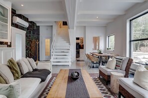 A modern living room with a sofa, coffee table, and a dining area with chairs and a table. A staircase leads to an upper level.