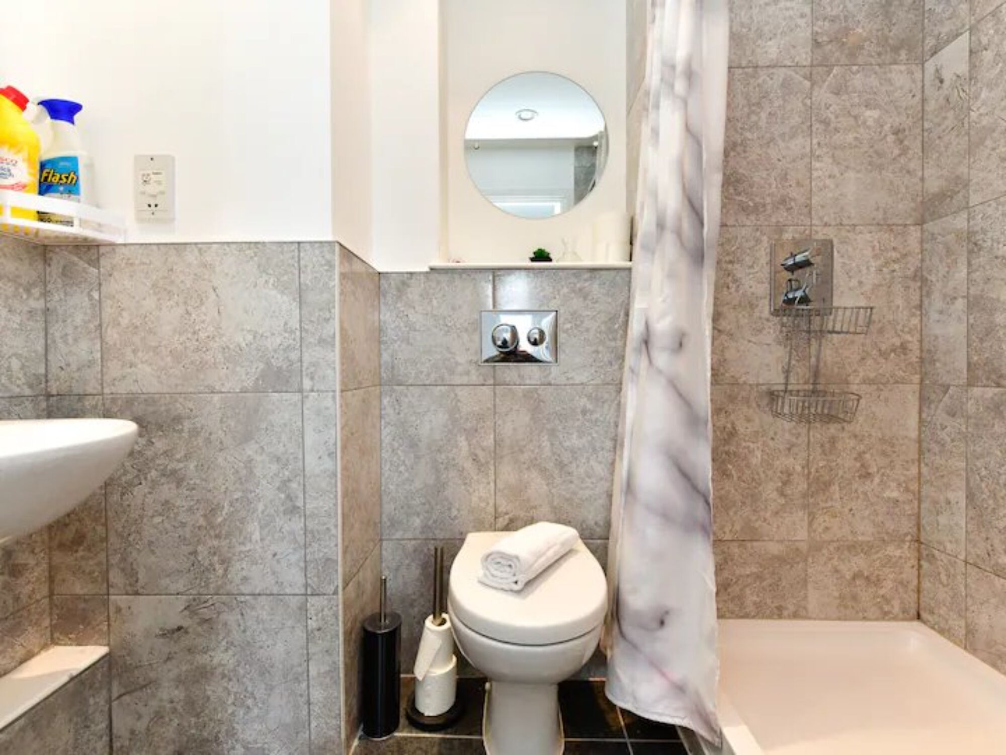 A bathroom with a toilet, sink, and shower. There is a mirror above the toilet, and a shelf with cleaning supplies.
