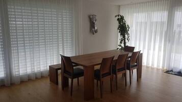 Ein Essbereich mit Holztisch und Stühlen, großen Fenstern mit durchsichtigen Vorhängen und einem Blumentopf.