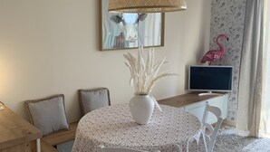 A dining area with a round table, white chairs, a woven pendant light, a vase with pampas grass, a framed picture, and a pink flamingo decoration.