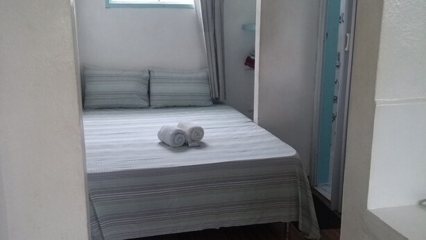 A small, narrow room with a single bed, a window with curtains, and a wooden floor. - Antonia Hospedaria (Búzios)