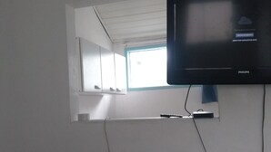 A television mounted on a white wall with a window above it, a power outlet below, and a visible crack on the wall. - Antonia Hospedaria (Búzios)
