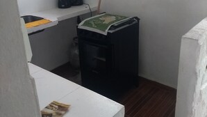 A small kitchen with a white countertop, a black trash bin, a coffee maker, and a window with curtains. - Antonia Hospedaria (Búzios)