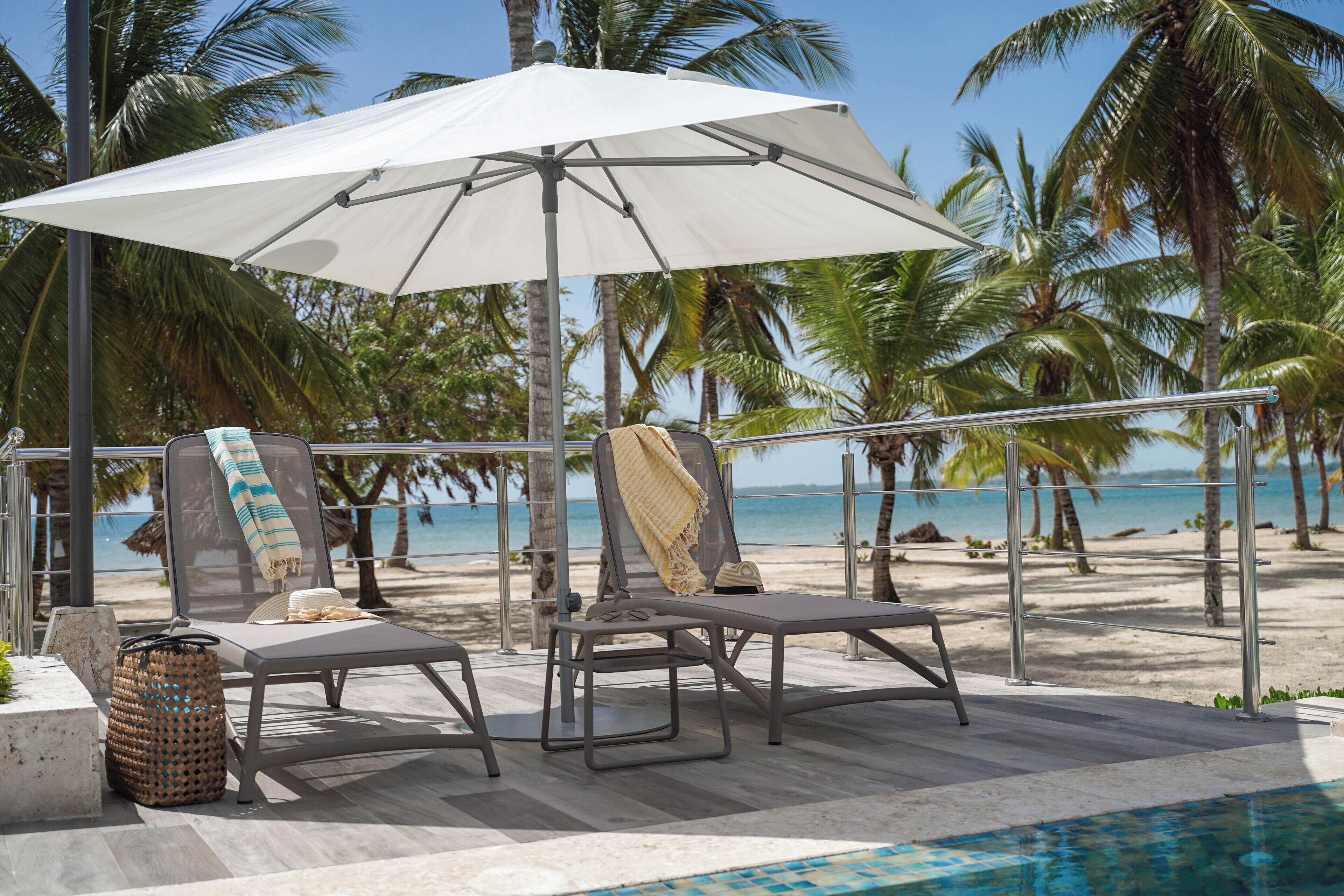 A poolside area with two lounge chairs, a white umbrella, and a view of the beach and palm trees.