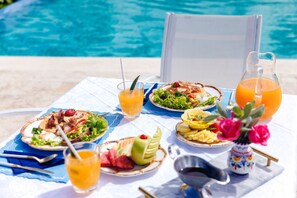 Una mesa dispuesta con diversos platos, bebidas y un jarrón de flores junto a una piscina.