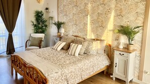 Une chambre à coucher avec un grand lit, une tête de lit en bois, une table de chevet, une commode et un mur orné d’un motif de feuilles.